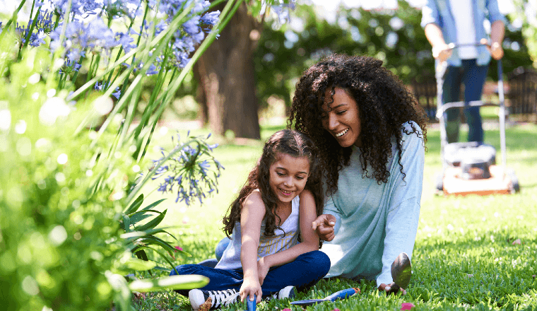 Mother and daughter sitting on the lawn with dad mowing lawn in the background